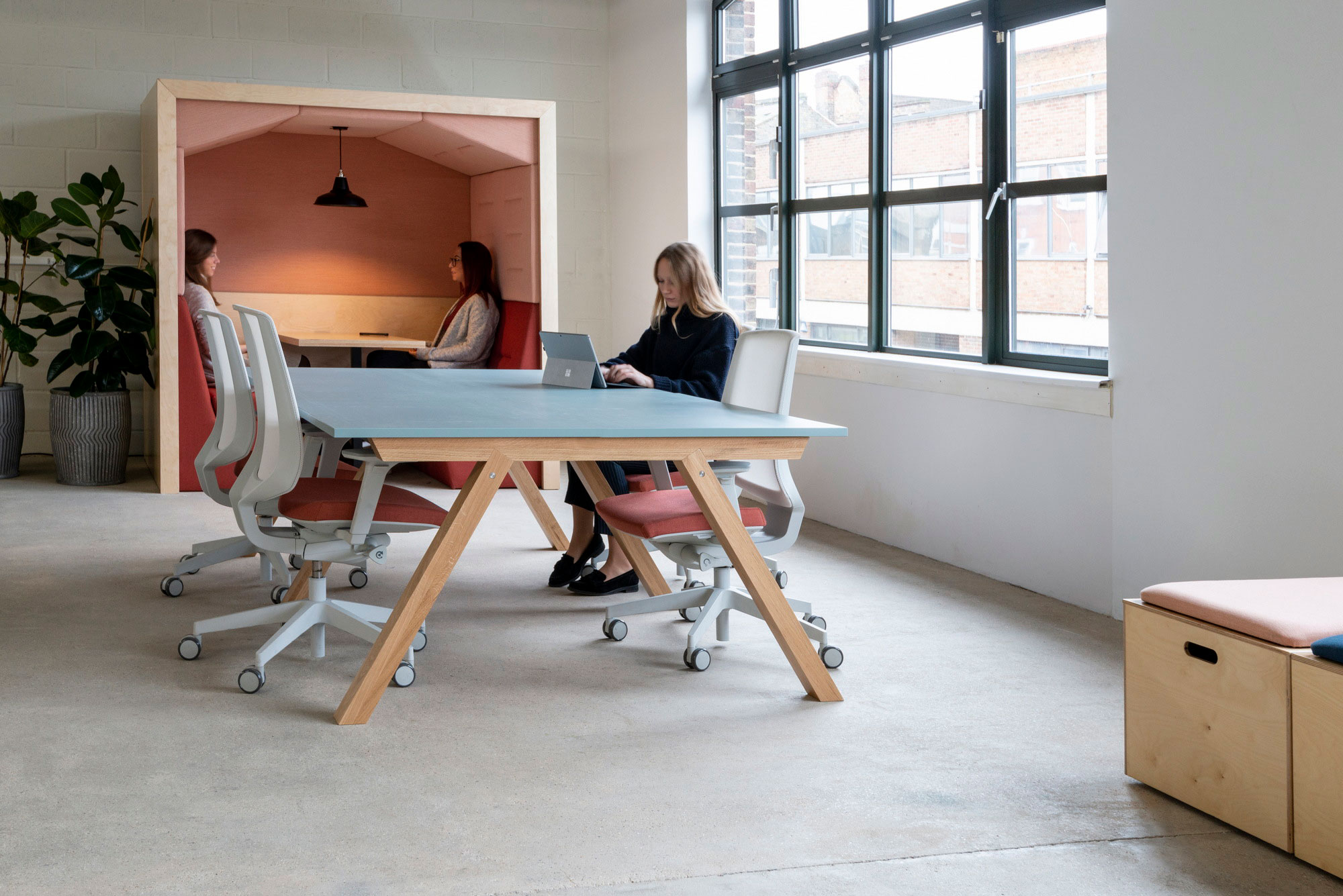 A meeting area with a pink acoustic booth and people working at a blue table beside tall windows
