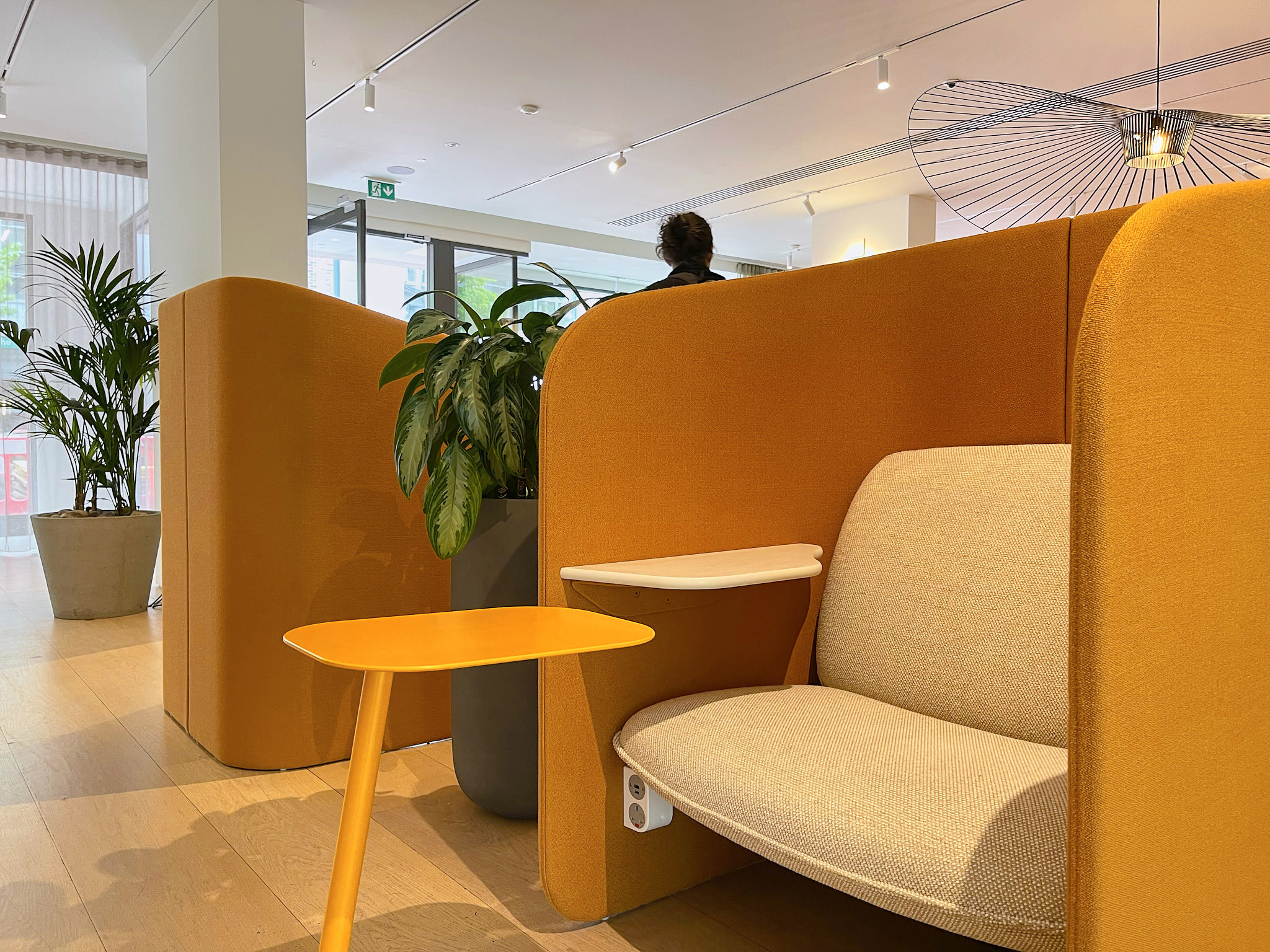 An orange acoustic booth with an upholstered bench seat and a small side table, beside planting in an office