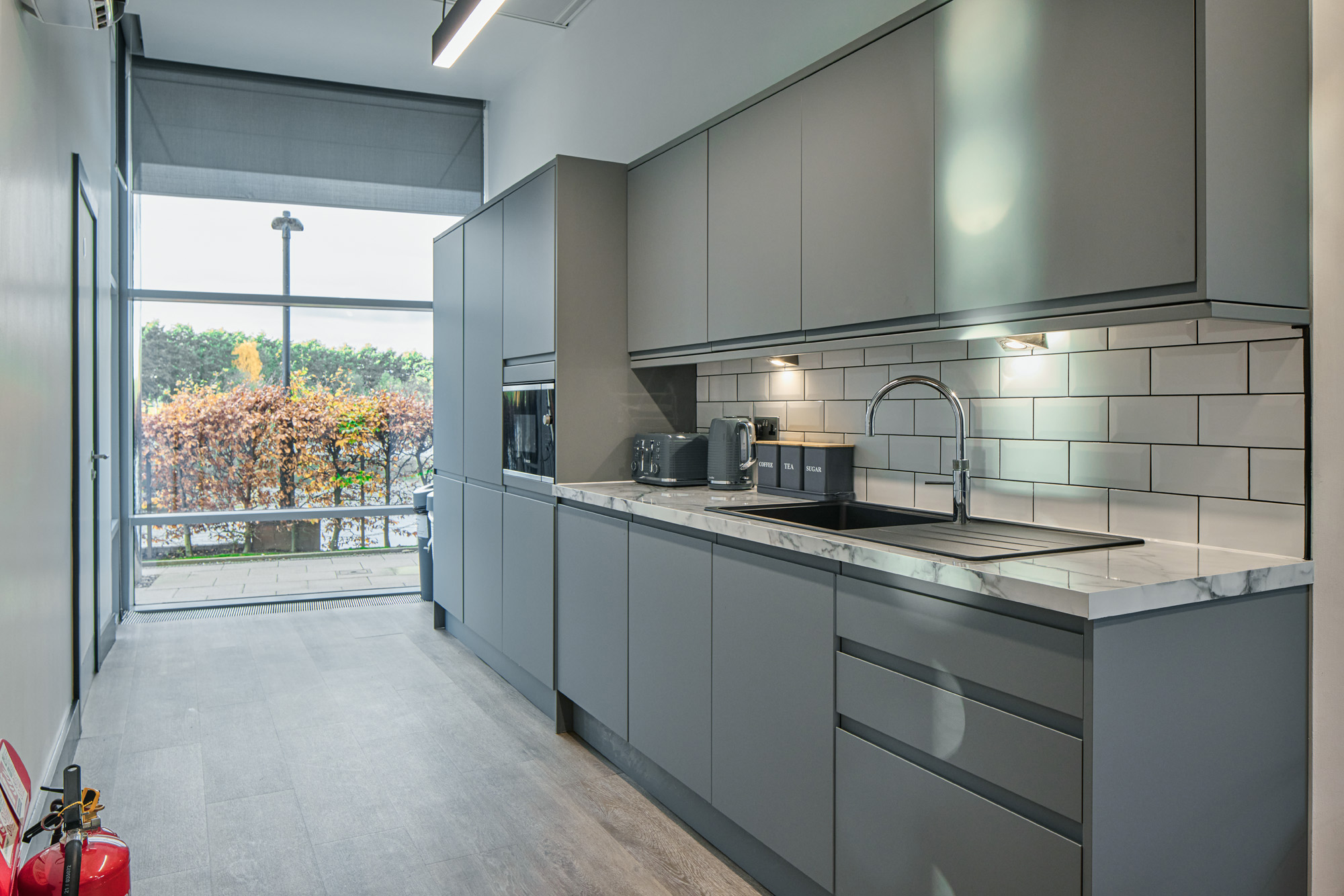 A fitted tea point in a London office, with grey units, a tiled splashback and a window onto planting
