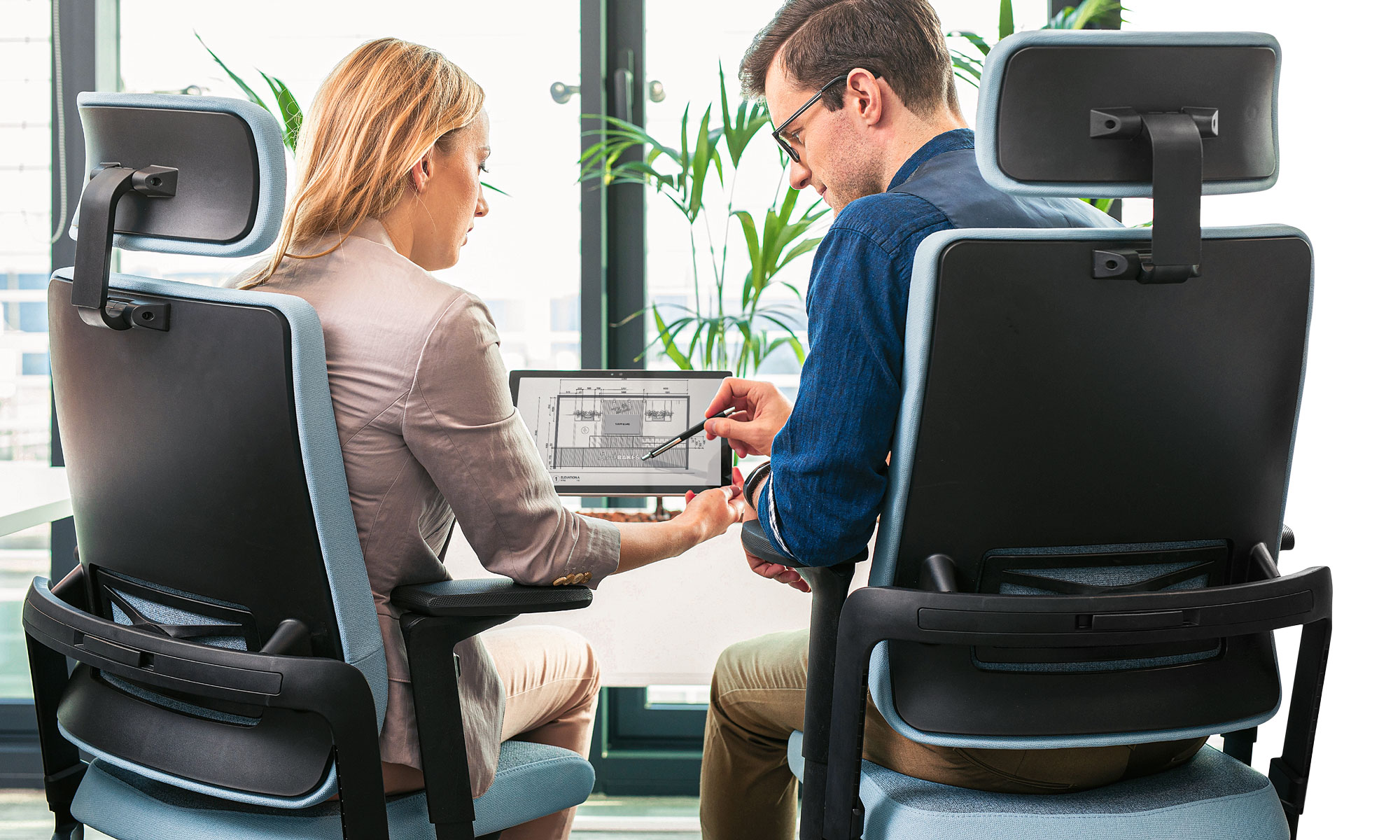 Two colleagues at a desk looking at a screen together, seated in high-backed ergonomic chairs