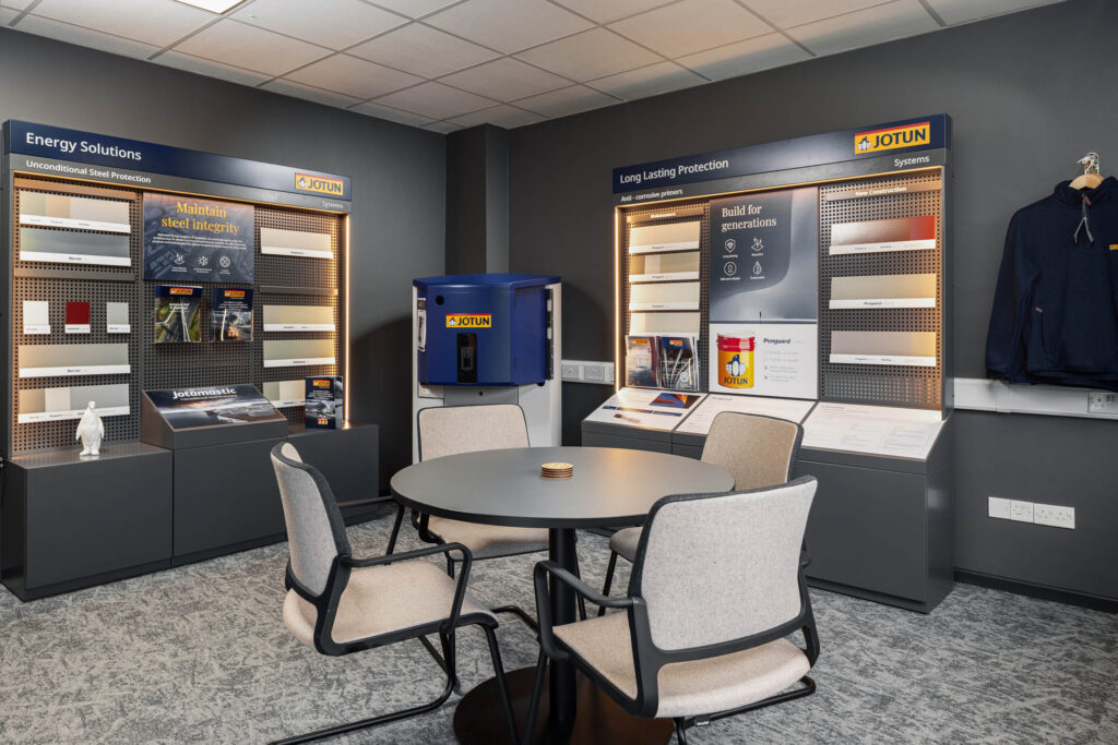 Modern showroom interior with Jotun product displays showcasing steel protection systems and coatings, featuring sample panels, branded literature, and a round meeting table with four upholstered chairs.